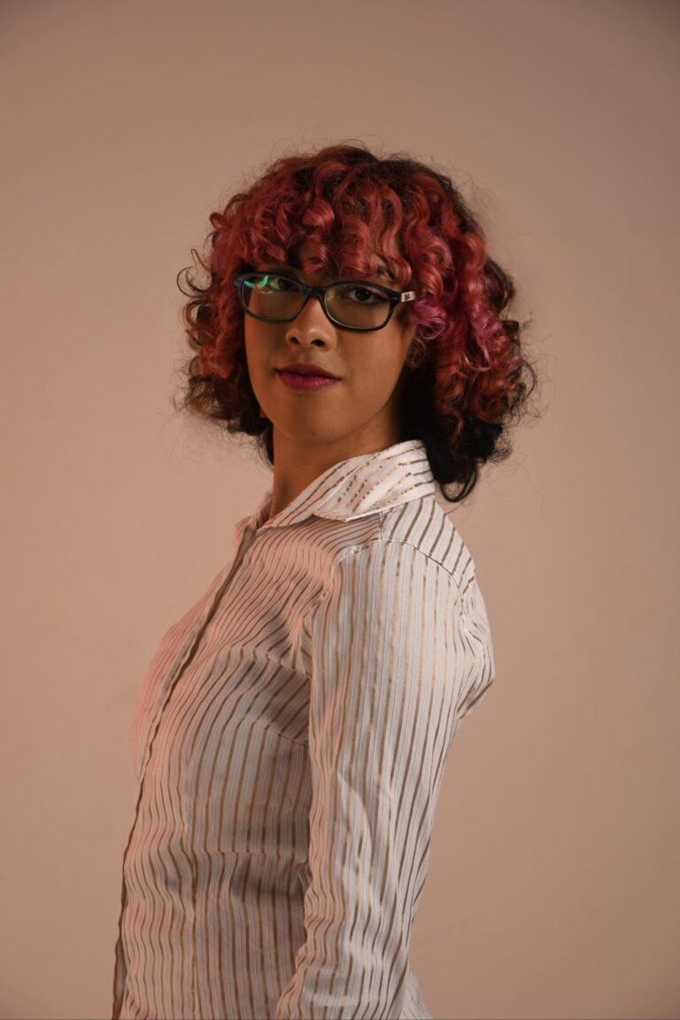 Sabrina Conconi, professora de japonês. Retrato de uma pessoa jovem de pele parda com cabelos cacheados, tingidos de rosa na parte superior e pretos na parte inferior. Ela usa óculos de armação preta e veste uma camisa clara com listras verticais finas e douradas. A pessoa está posicionada de lado, mas vira o rosto para olhar diretamente para a câmera com uma expressão neutra. O fundo é bege liso.
